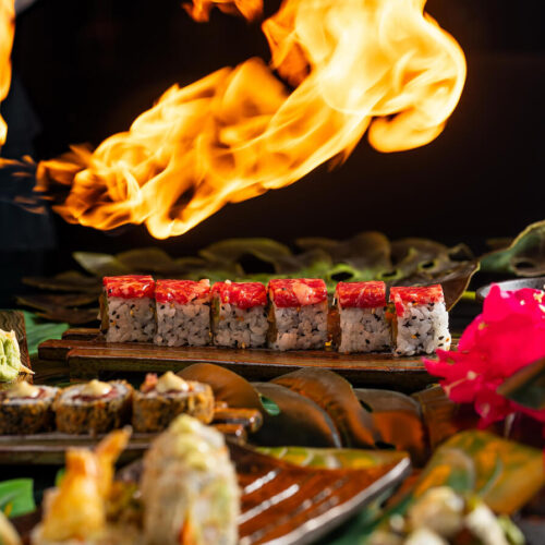 Flame-seared sushi rolls topped with tuna, set against a dramatic fire backdrop and surrounded by tropical decor
