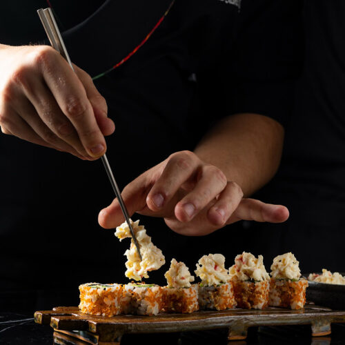 Chef carefully plating sushi rolls with creamy topping using metal chopsticks, showcasing Japanese culinary precision.